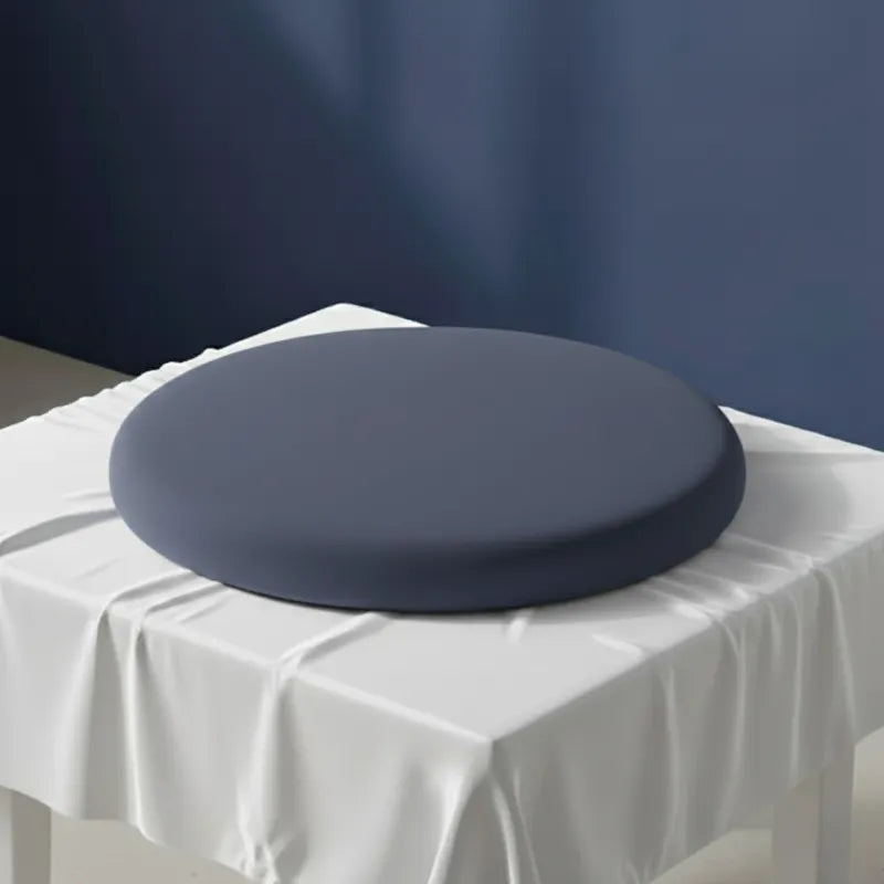Coussin ergonomique rond bleu foncé pour chaise de bureau, en mousse mémoire de forme, sur un drap satiné gris perle avec mur bleu en arrière-plan.