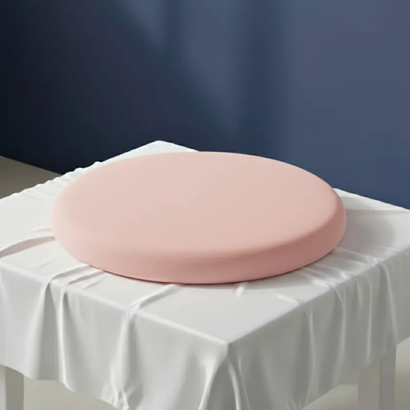 Coussin ergonomique rond rose pour chaise de bureau, en mousse mémoire de forme. Posé sur une table recouverte d’un drap satiné gris perle, devant un mur bleu profond à la lumière naturelle.