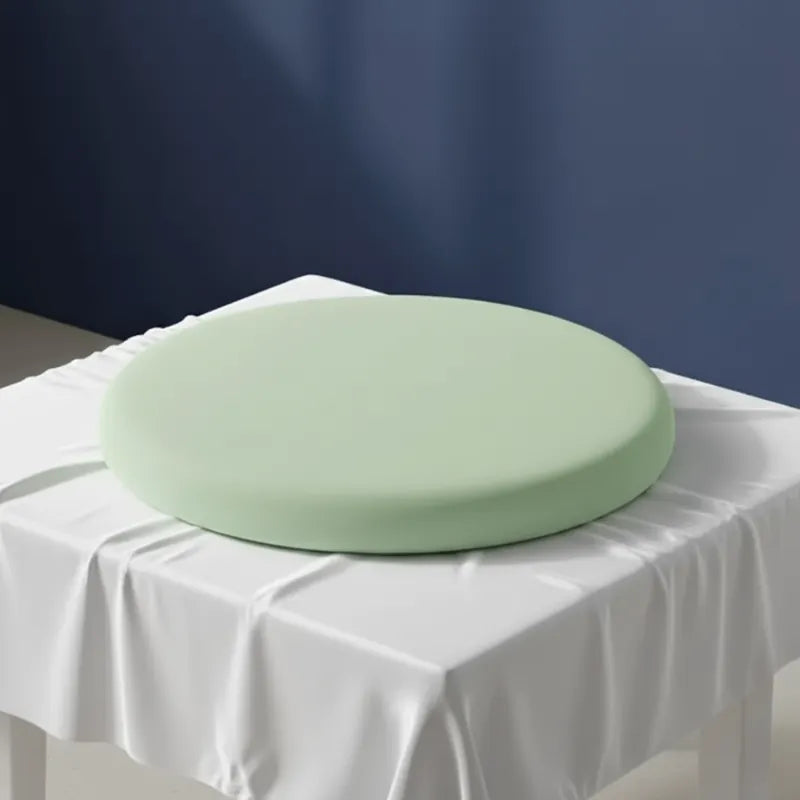 Coussin ergonomique rond vert clair pour chaise de bureau, en mousse mémoire de forme, sur une table recouverte d’un drap satiné gris perle devant un mur bleu.