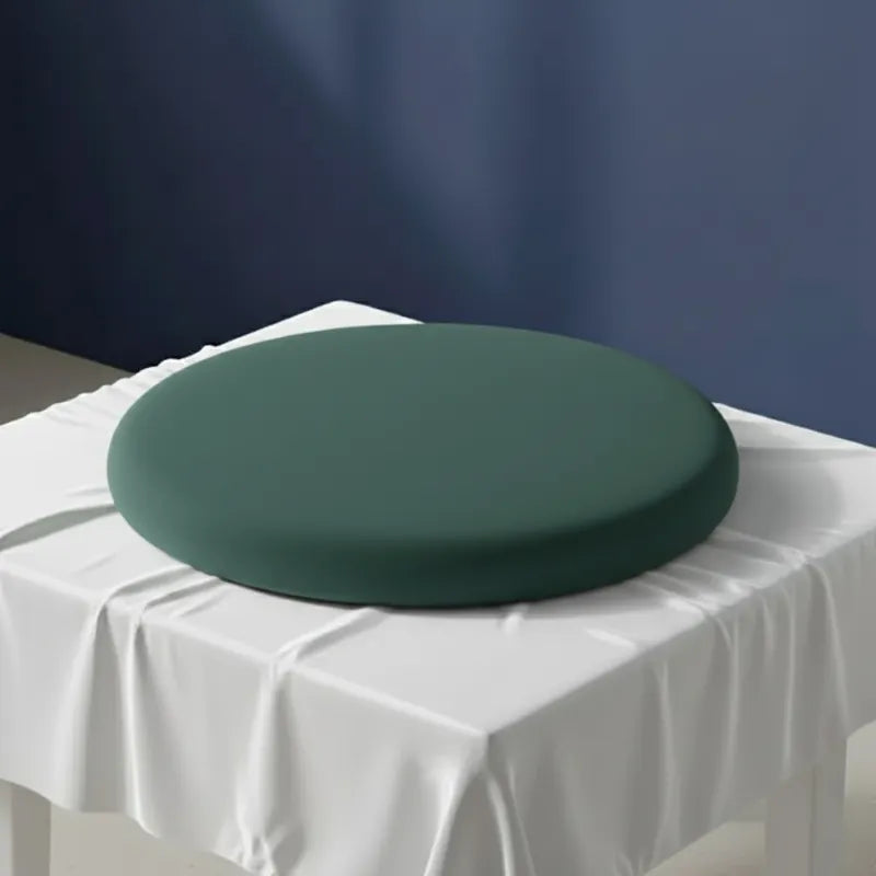Coussin ergonomique rond vert foncé pour chaise de bureau, en mousse mémoire de forme, posé sur un drap satiné gris perle devant un mur bleu.