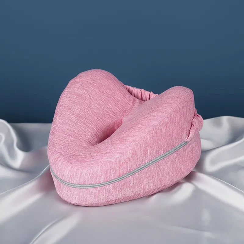 Coussin ergonomique entre les jambes rose sur drap gris perle, fond bleu uni et éclairage doux