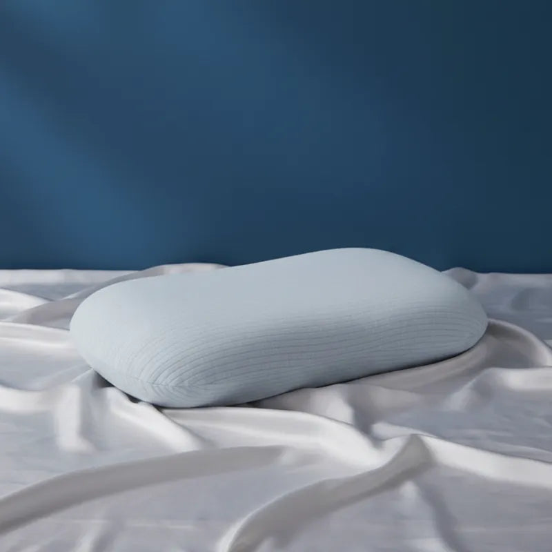 oreiller ergonomique en latex naturel bleu entièrement recouvert, sur drap satiné gris perle et mur bleu profond, éclairage doux naturel, rendu haut de gamme