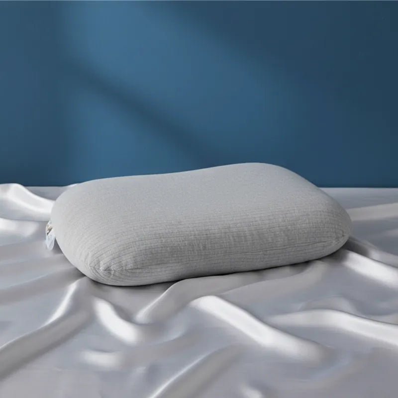 oreiller ergonomique en latex naturel gris entièrement recouvert, posé sur drap satiné gris perle et mur bleu profond, lumière naturelle douce