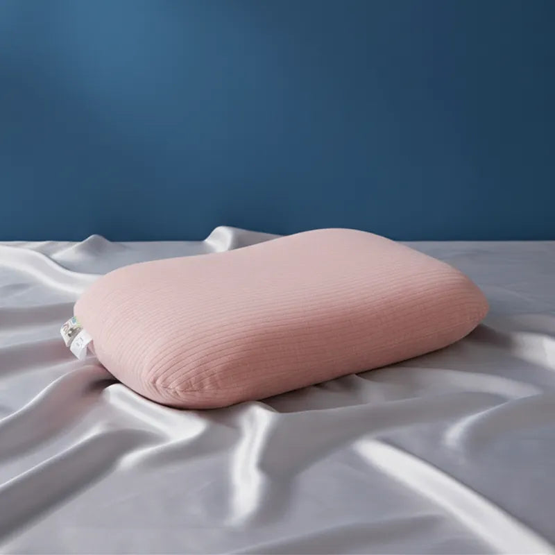 oreiller ergonomique en latex naturel rose entièrement recouvert, sur drap satiné gris perle avec mur bleu profond et éclairage naturel doux