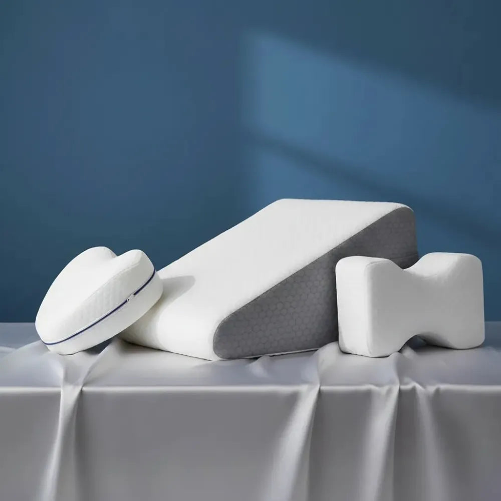 Coussins ergonomiques en mémoire de forme pour les jambes — formes orthopédiques variées pour soutenir les jambes et les genoux, présentés sur un drap satiné gris perle devant un mur bleu profond.