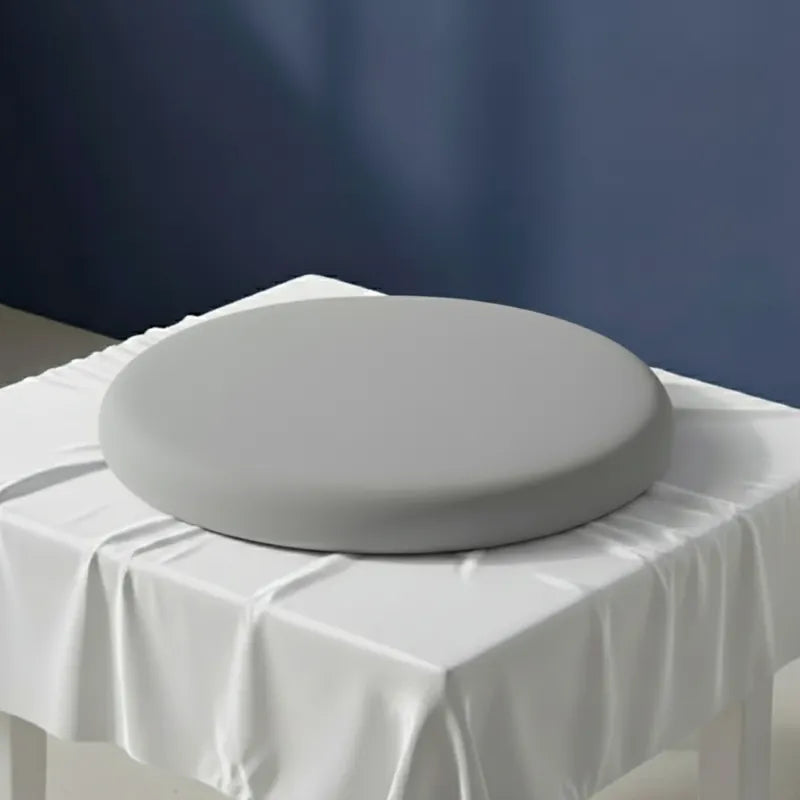 Coussin ergonomique rond gris pour chaise de bureau, en mousse mémoire de forme, posé sur un drap satiné gris perle avec fond bleu.