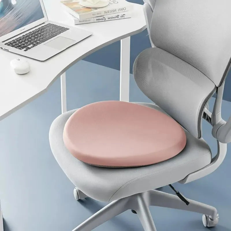 Coussin ergonomique rond rose sur une chaise de bureau grise, devant un bureau moderne avec ordinateur portable. Accessoire de confort pour une assise prolongée.