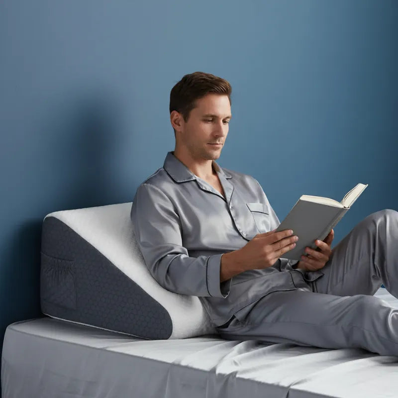 Homme européen en pyjama satiné lisant appuyé sur un oreiller ergonomique carré 50x50 blanc et gris, fond bleu profond